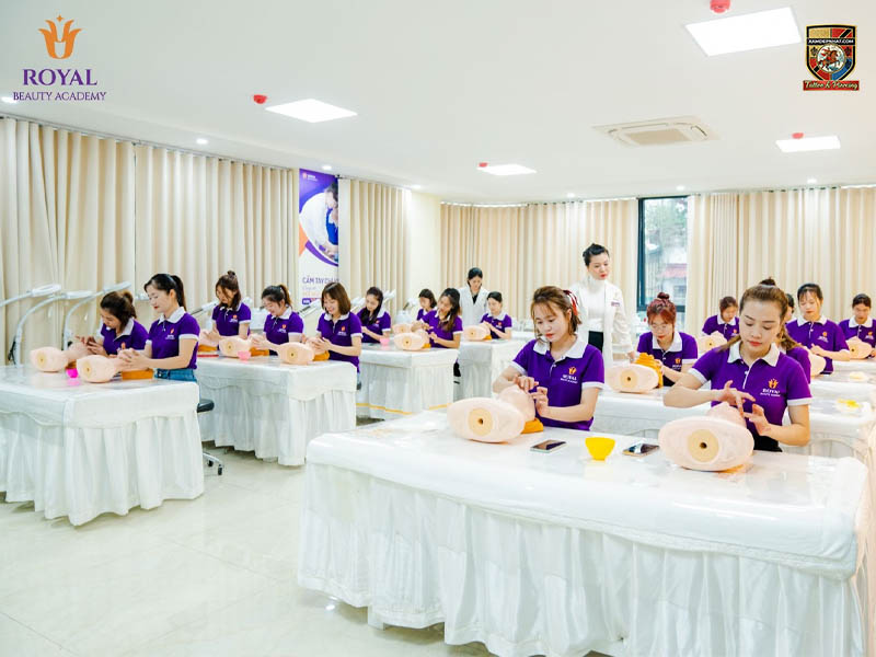 Học viên Royal Beauty Academy mặc đồng phục tím đang thực hành chăm sóc da trên ma nơ canh dưới sự hướng dẫn của giảng viên.