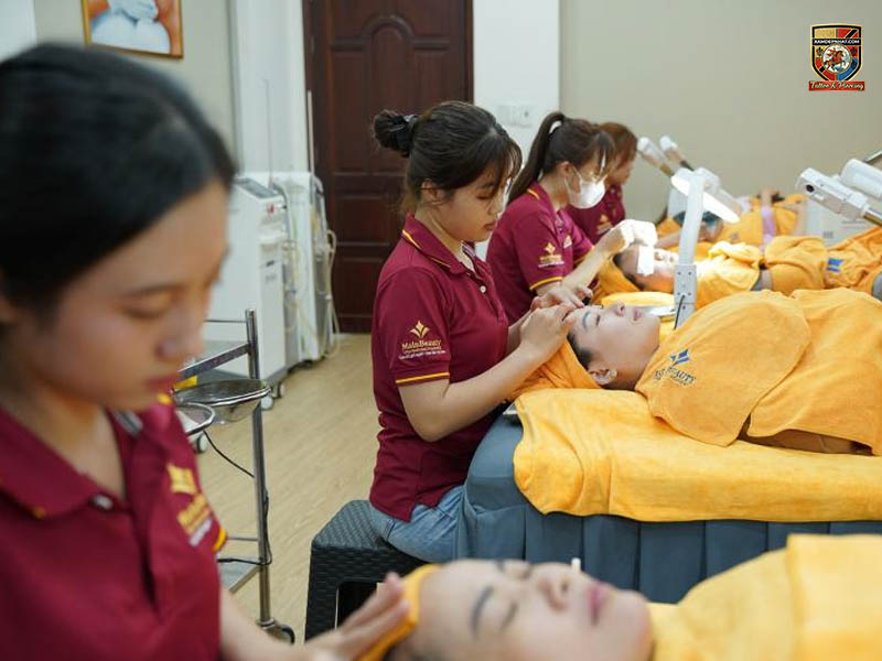 Học viên MainBeauty mặc đồng phục đỏ đô đang thực hành các kỹ thuật chăm sóc da mặt và massage cho khách hàng tại lớp học spa.