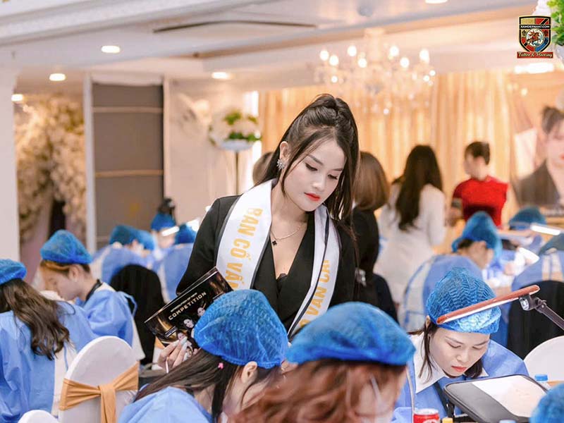 Ban cố vấn cuộc thi phun xăm thẩm mỹ đang quan sát và chấm thi các thí sinh trong hội trường lớn.