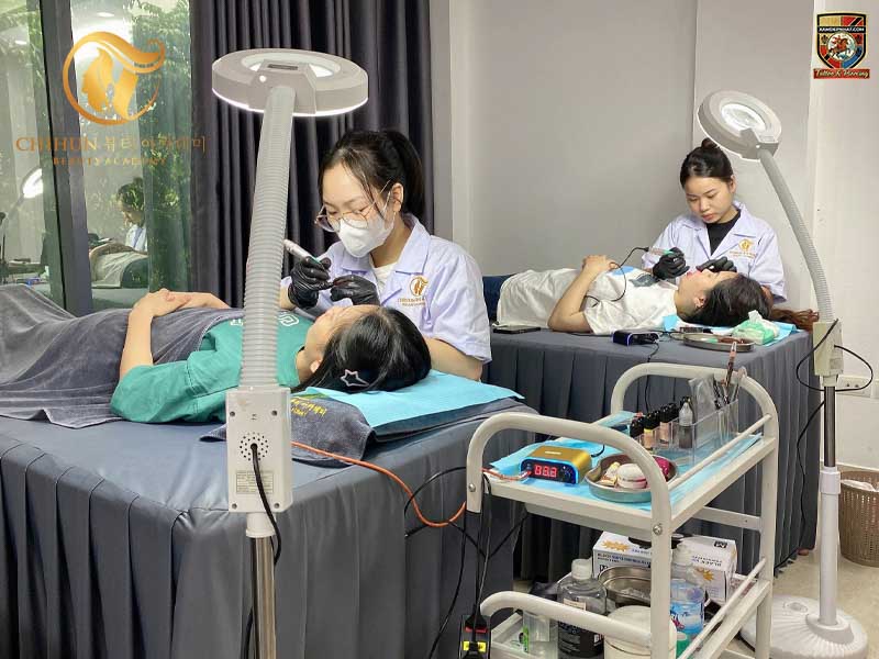 Ảnh chụp hai kỹ thuật viên đang tập trung thực hành kỹ thuật phun xăm thẩm mỹ chân mày trên hai khách hàng khác nhau tại một học viện làm đẹp.