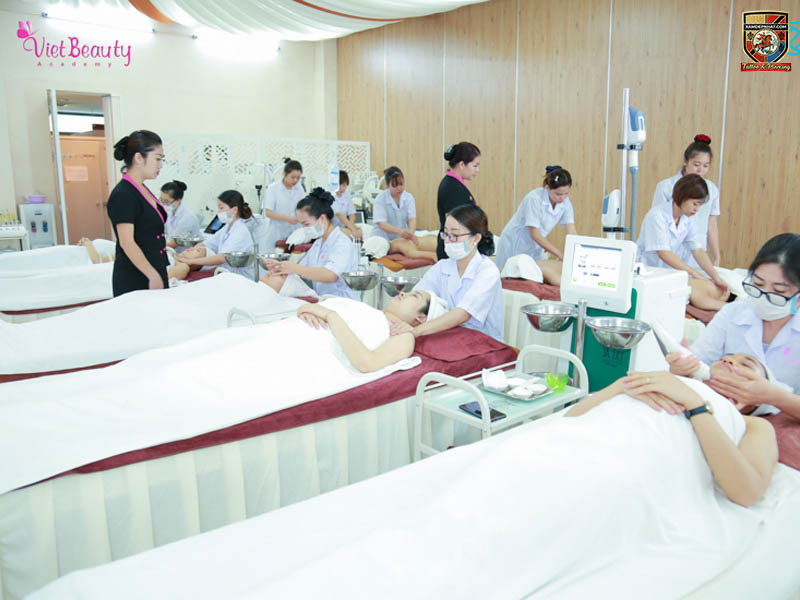 Lớp học thực hành tại Học viện VietBeauty Academy, với nhiều học viên đang chăm sóc da mặt và thực hiện các kỹ thuật mát-xa trên người mẫu nằm trên giường.