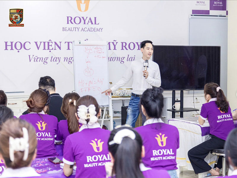 Male instructor lecturing on technical formulas on a whiteboard to students wearing purple Royal Beauty Academy uniforms
