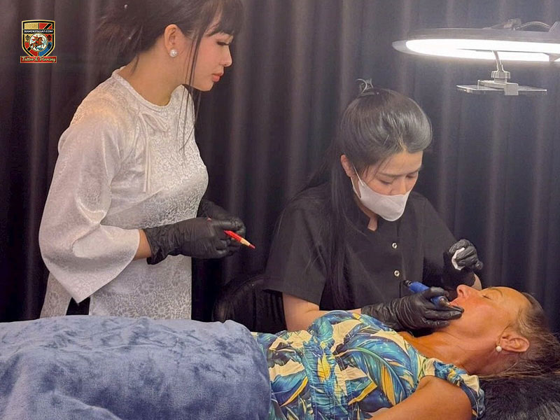 Senior master in traditional attire supervising a student performing precision cosmetic tattooing on a client