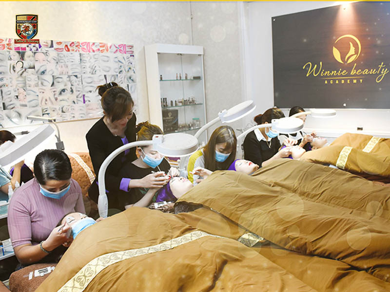 Beauty academy practical room with multiple beds and ring lamps where students practice PMU and lash techniques