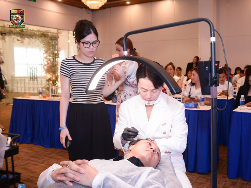 Chuyên gia mặc vest trắng thao tác tạo hình chân tóc dưới đèn vòm trước sự quan sát của học viên