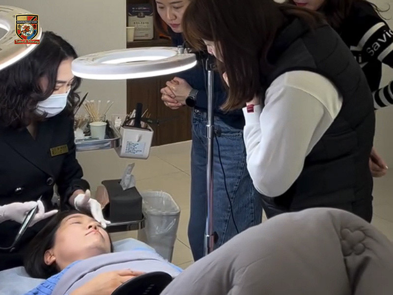 Chuyên gia đeo găng tay trắng thực hiện kỹ thuật chân mày và viền tóc dưới đèn led tròn cho học viên quan sát