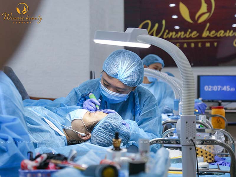 Trainees in surgical gowns and masks practicing under bright studio lighting at Winnie Beauty Academy